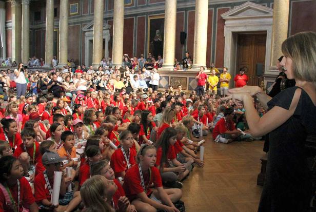 Eine Frau spricht vor einer großen Gruppe von Kindern in einem historischen Gebäude.