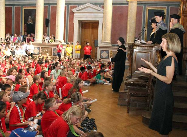 Eine Frau spricht vor einer Gruppe von Kindern in einem großen Saal.