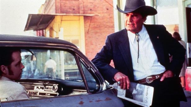 Ein Mann mit Cowboyhut lehnt an einem Auto und hält eine Zeitung in der Hand.
