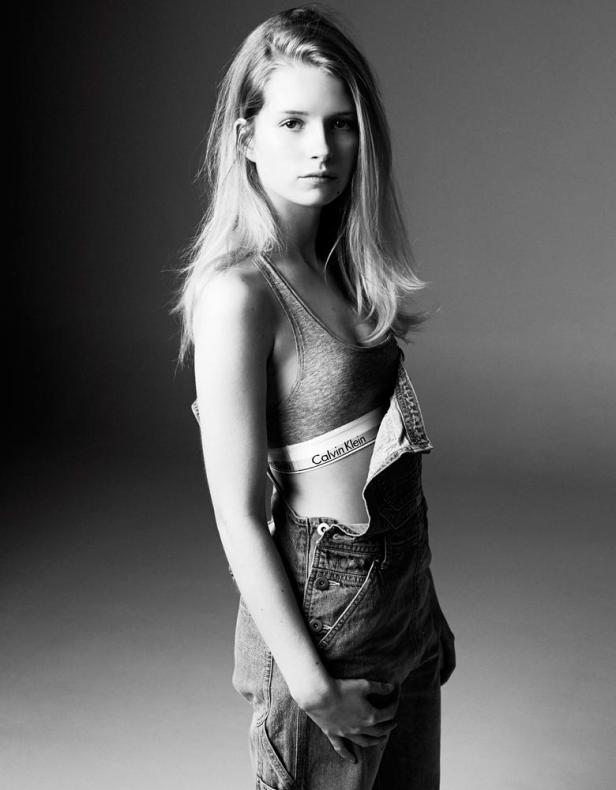 Eine junge Frau in Jeanslatzhose und einem Calvin Klein Sport-BH posiert in Schwarzweiß.