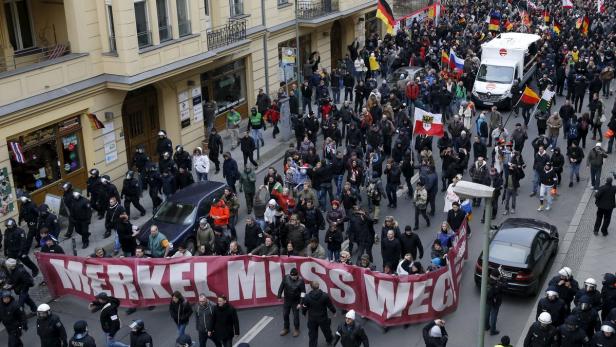 Eine Menschenmenge demonstriert mit einem Banner „Merkel muss weg“ in einer Stadt.