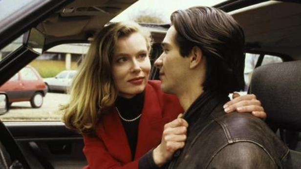 Eine Frau im roten Blazer berührt den Mann im Auto zärtlich an der Schulter.