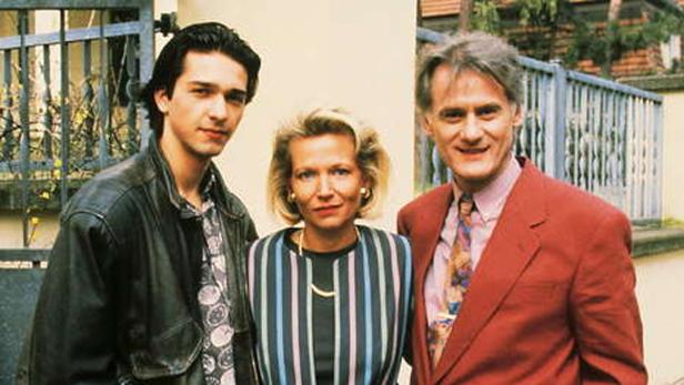 Drei Personen posieren vor einem Haus, ein junger Mann mit Lederjacke, eine Frau und ein Mann im roten Anzug.