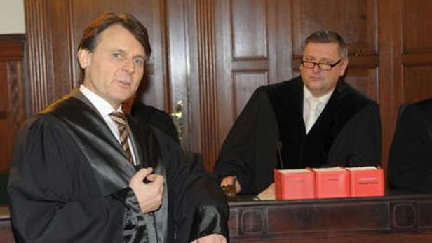 Zwei Richter in Roben sitzen an einem Richtertisch mit roten Aktenordnern.