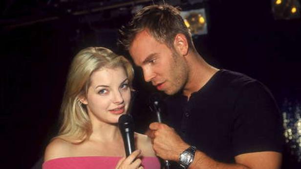 Blonde Frau und Mann mit Mikrofonen auf einer Bühne.