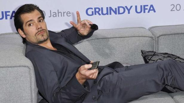 Ein Mann im Anzug liegt auf einem grauen Sofa und hält eine Fernbedienung in der Hand.