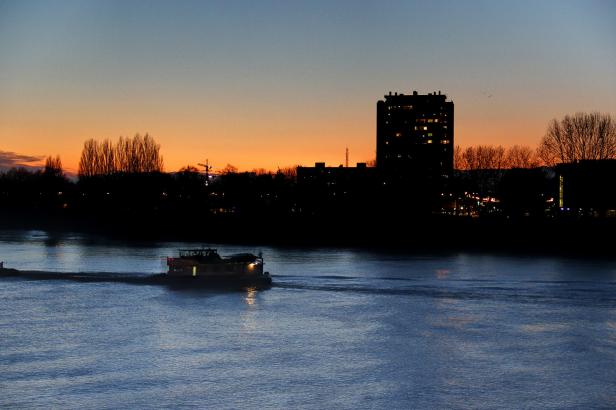 Ein Schiff fährt auf einem Fluss vor einer Silhouette aus Gebäuden im Abendlicht.