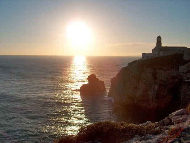 Sonnenuntergang über dem Meer mit einem Leuchtturm auf einer Klippe.