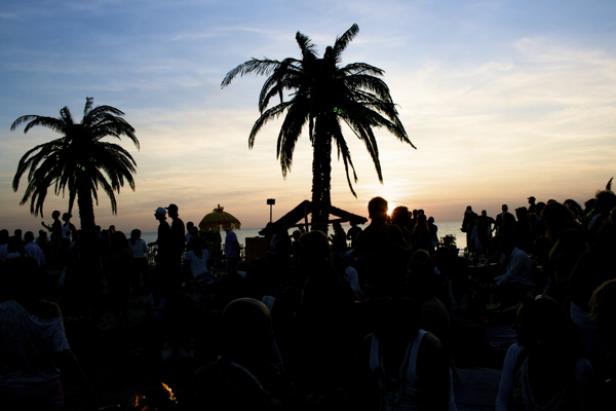 Im Gegenlicht stehen Palmen und eine Menschenmenge am Strand.
