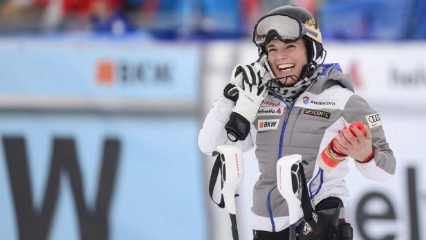 Eine lachende Skirennläuferin mit Helm und Skibrille hält ihre Skistöcke.