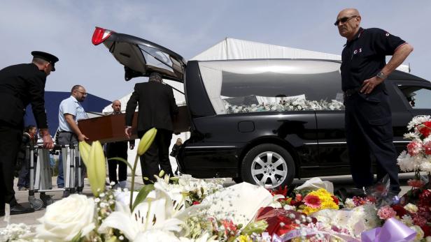 Ein Sarg wird in einen Leichenwagen gehoben, umgeben von Blumen.