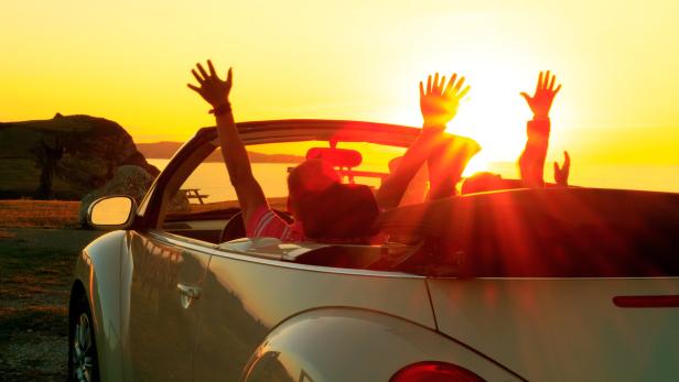 Menschen fahren in einem Cabrio bei Sonnenuntergang und heben die Arme.