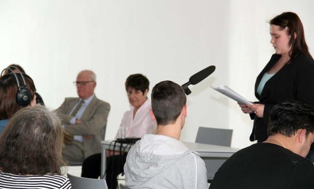 Eine Frau liest vor Publikum aus einem Dokument vor.