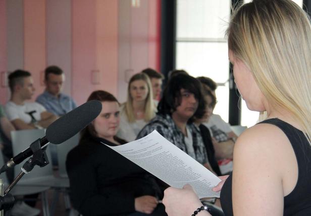 Eine junge Frau liest vor einer Gruppe von Zuhörern aus einem Blatt Papier vor.