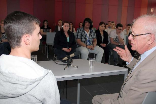 Ein älterer Mann im Gespräch mit einem jungen Mann vor Publikum.