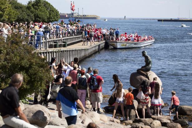 Die Kleine Meerjungfrau in Kopenhagen wird von Touristen besucht, während ein Boot mit Weihnachtsmännern vorbeifährt.