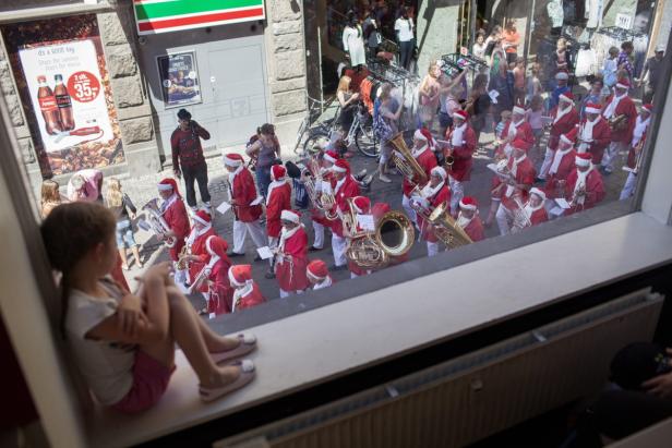 Ein Mädchen sitzt am Fenster und beobachtet eine Blaskapelle in Weihnachtsmannkostümen.