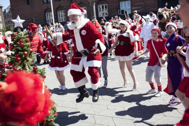 Eine Gruppe von Menschen, darunter mehrere Weihnachtsmänner und -frauen, tanzen auf einem Platz.