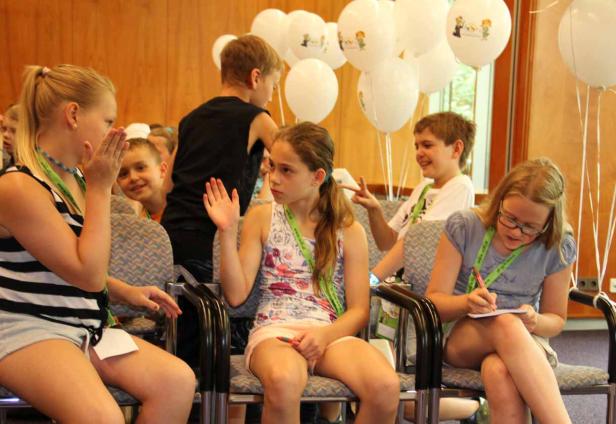 Kinder sitzen auf Stühlen und scheinen an einer Aktivität teilzunehmen; Ballons sind im Hintergrund.