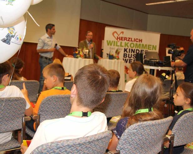 Kinder sitzen bei einer Veranstaltung vor einer Bühne mit Rednern.