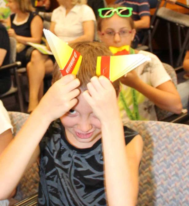 Ein Junge hält gelb-rote Papierhüte über seinen Kopf, während ein anderes Kind im Hintergrund sitzt.