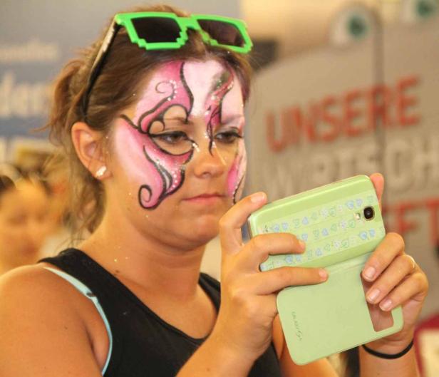 Eine Frau mit Gesichtsbemalung und Pixel-Sonnenbrille fotografiert mit ihrem Smartphone.