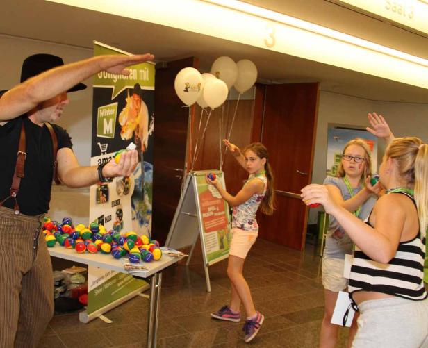 Ein Jongleur mit Hut und Hosenträgern jongliert mit Kindern mit bunten Bällen.