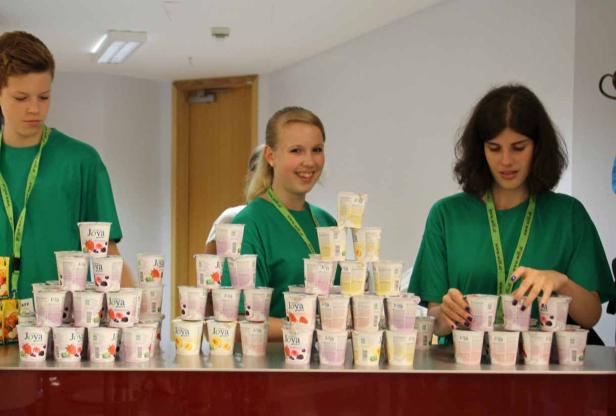 Drei Personen in grünen T-Shirts stapeln Joghurtbecher der Marke Joya zu Pyramiden.