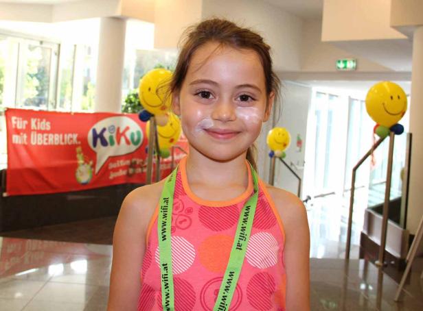 Ein Mädchen mit Sonnencreme im Gesicht lächelt in die Kamera. Im Hintergrund sind Ballons und ein Kiku-Banner zu sehen.