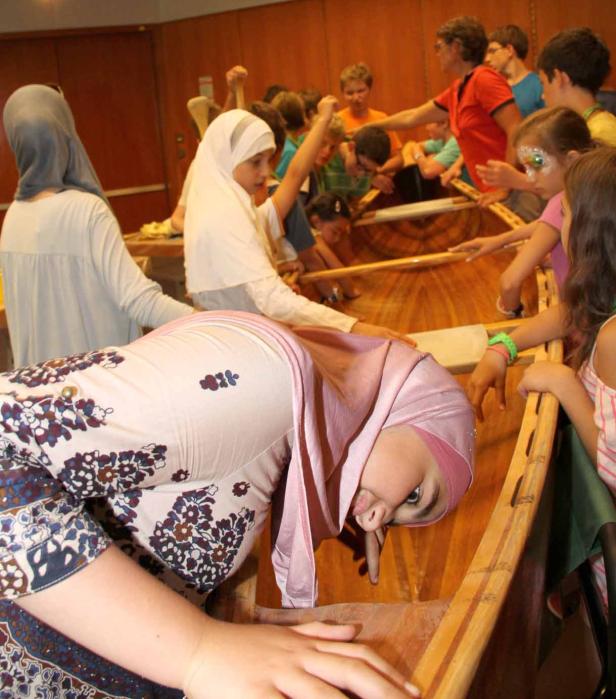 Eine Gruppe von Kindern, einige mit Hijabs, hantiert mit einem Holzboot in einem Innenraum.