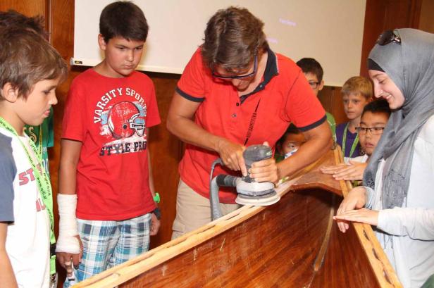 Eine Frau schleift ein Holzboot, umgeben von einer Gruppe interessierter Kinder.