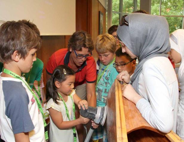 Eine Gruppe Kinder und ein Erwachsener arbeiten an einem Holzboot mit einem Schleifgerät.