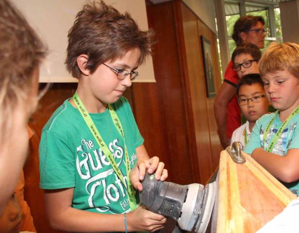 Ein Junge mit Brille schleift Holz mit einem elektrischen Schleifgerät.