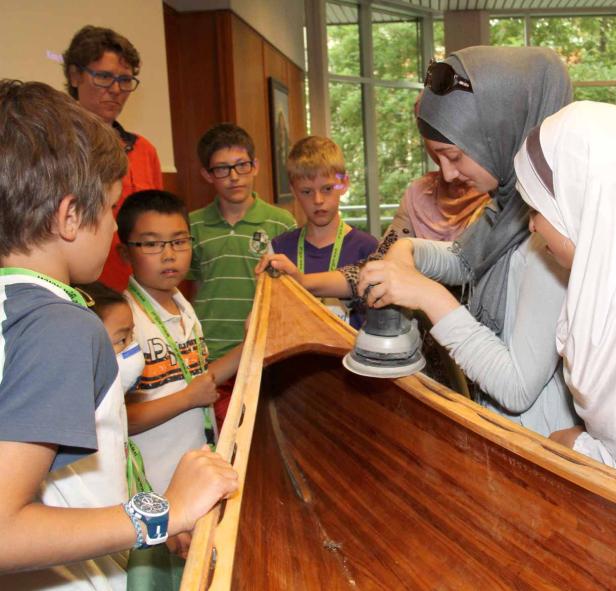 Eine Frau schleift mit einer Schleifmaschine ein Holzboot, umgeben von Kindern.