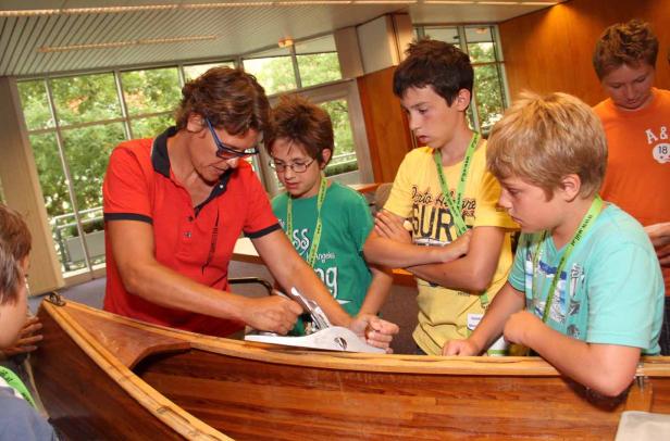 Ein Mann zeigt einer Gruppe von Kindern, wie man ein Holzboot hobelt.