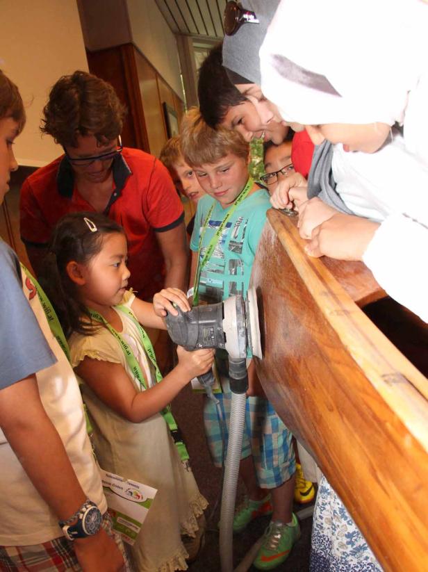 Ein junges Mädchen schleift ein Holzstück mit einem elektrischen Schleifer, umgeben von anderen Kindern und einem Erwachsenen.