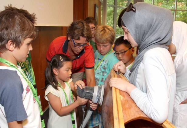 Eine Gruppe Kinder lernt unter Anleitung, wie man ein Holzboot schleift.