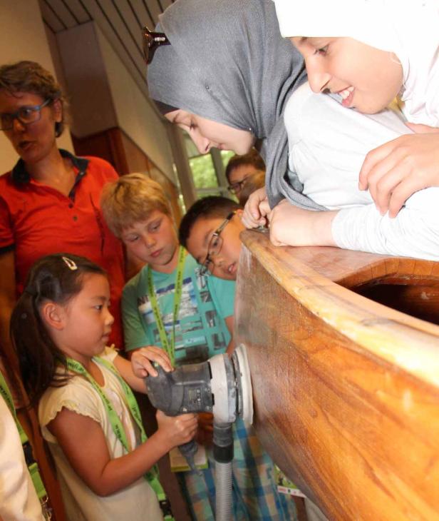 Eine Gruppe von Kindern schleift unter Aufsicht ein Holzboot.