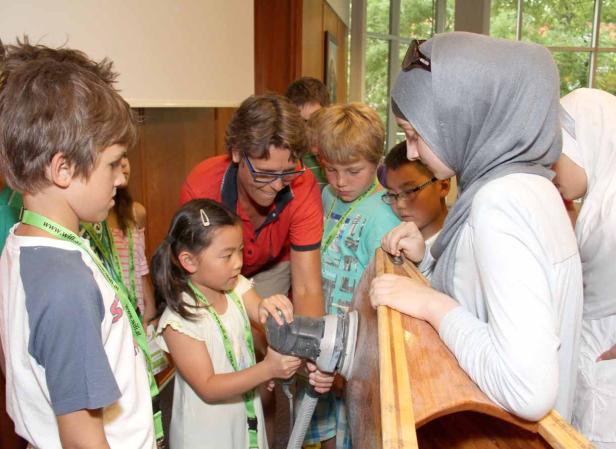 Eine Gruppe von Kindern und ein Erwachsener arbeiten an einem Holzboot mit einem Schleifgerät.