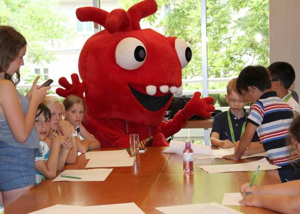 Eine Gruppe Kinder sitzt mit einem großen, roten Maskottchen an einem Tisch und malt.