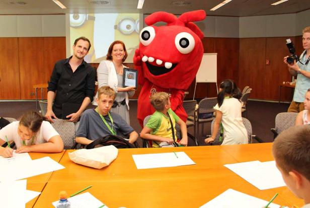 Kinder zeichnen an einem Tisch, während ein rotes Maskottchen und Erwachsene zusehen.