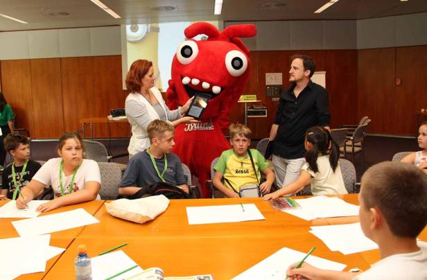 Kinder sitzen an einem Tisch und zeichnen, während eine Frau und ein Maskottchen einen Bildschirm zeigen.
