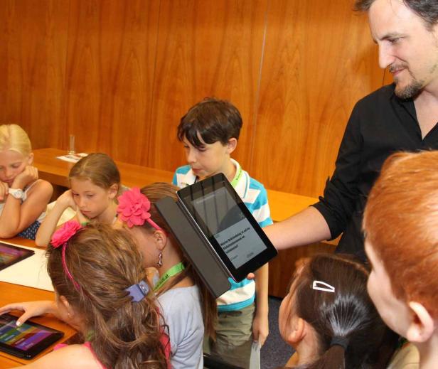 Eine Gruppe Kinder sitzt an einem Tisch und schaut auf Tablets, die von einem Mann präsentiert werden.