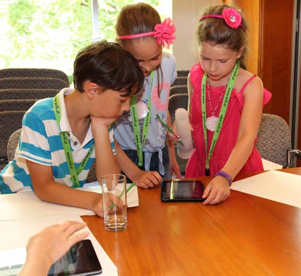 Drei Kinder stehen an einem Tisch und schauen auf ein Tablet.