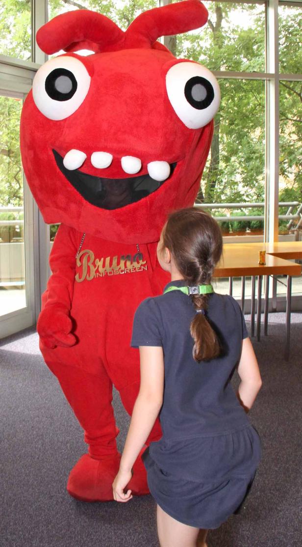 Ein Mädchen steht vor einem großen, roten Maskottchen namens Bruno Infoscreen.