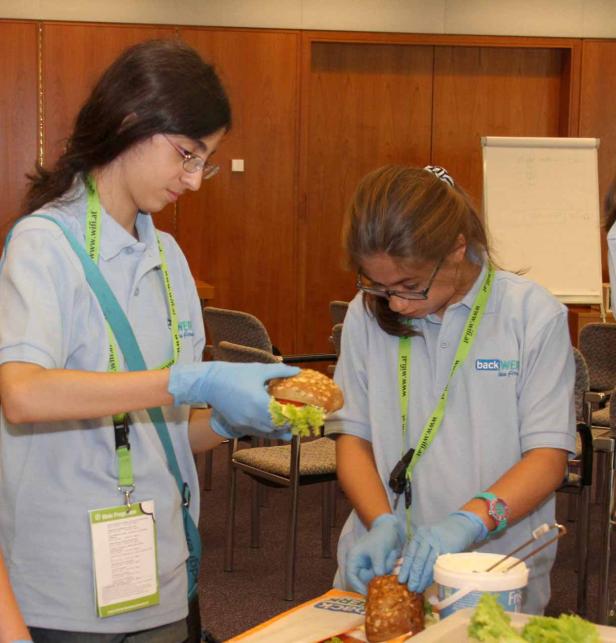 Zwei junge Frauen bereiten mit Handschuhen belegte Brötchen zu.