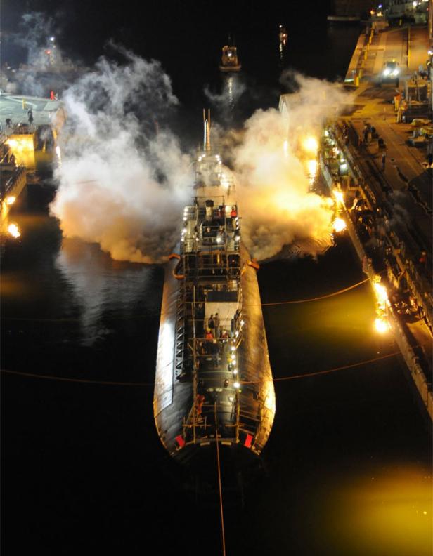 Ein U-Boot wird nachts in einem Hafenbecken zu Wasser gelassen, begleitet von Rauch und Lichtern.