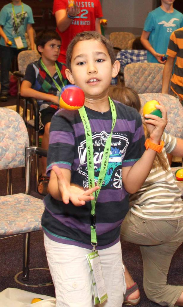 Ein Junge jongliert mit drei Bällen in einem Raum voller Menschen.