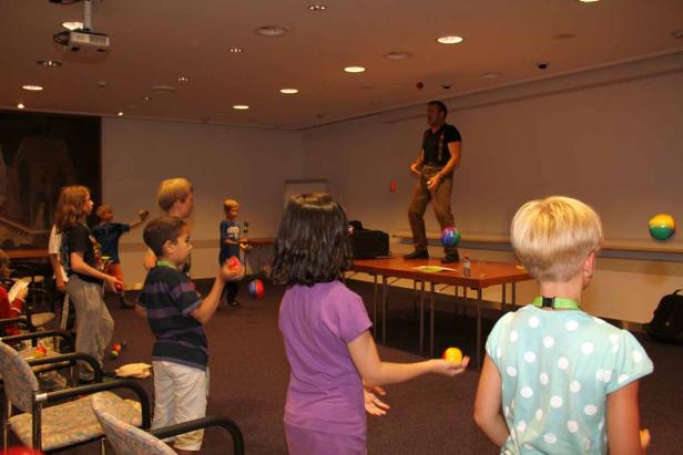 Ein Mann auf einem Tisch jongliert mit Bällen vor einer Gruppe von Kindern.