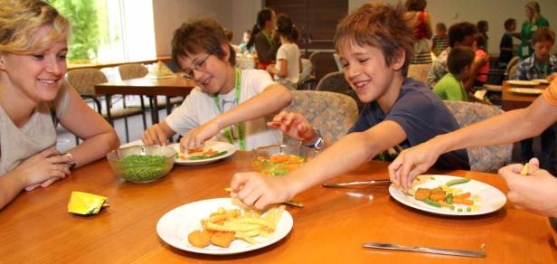 Kinder und eine Frau sitzen an einem Tisch und belegen ihre Teller mit Gemüse und Pommes.